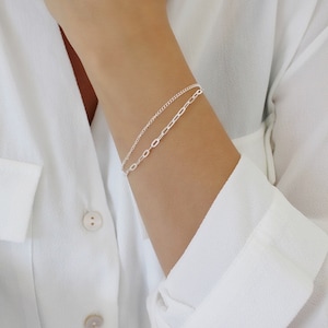 May include: Two silver chain bracelets layered on a wrist. The top bracelet is a delicate chain, while the bottom bracelet is a thicker chain with oval links.
