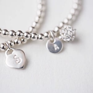 May include: Two silver bracelets with charms. One charm is a heart with the letter 'S' on it. The other charm is a round disc with the letter 'A' on it. The bracelets are made of small silver beads.