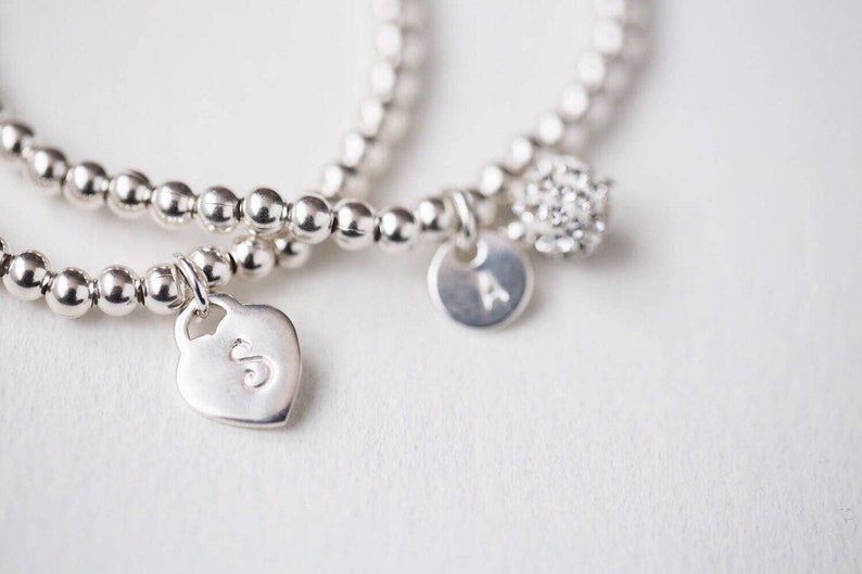 May include: Two silver bracelets with charms. One charm is a heart with the letter 'S' on it. The other charm is a round disc with the letter 'A' on it. The bracelets are made of small silver beads.