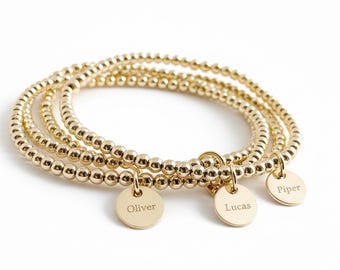 May include: A set of three gold-toned beaded bracelets, each featuring a small, round, flat charm. The charms are engraved with the names Oliver, Lucas, and Piper, respectively. The bracelets are constructed from small, uniform, reflective gold spheres.