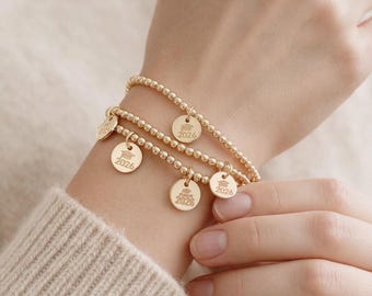May include: A set of three gold-toned beaded stretch bracelets, each featuring a small circular charm engraved with a graduation cap icon and the text 2026. The bracelets are composed of uniform, polished spherical beads arranged in a continuous loop.