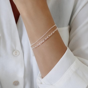 May include: A double-strand silver bracelet featuring one delicate fine curb chain and one slightly thicker elongated oval link chain. The two metallic silver chains are worn together as a single layered piece of jewellery on the wrist.