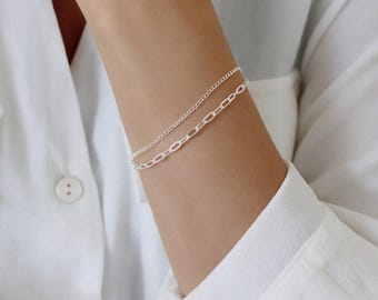May include: A double-strand silver bracelet featuring one delicate fine curb chain and one slightly thicker elongated oval link chain. The two metallic silver chains are worn together as a single layered piece of jewelry on the wrist.