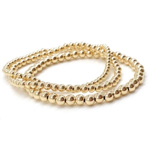 May include: Three gold beaded bracelets stacked on top of each other. The bracelets are made of small, round beads.