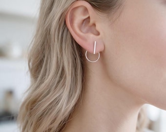 May include: A silver-toned earring featuring a thin vertical bar attached to a circular hoop. The earring is worn in the earlobe, with the straight bar positioned vertically above the circular hoop component, creating a geometric design.