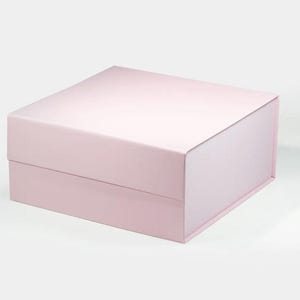Può includere: Una scatola regalo rettangolare di colore rosa pallido con un coperchio incernierato. La scatola è realizzata in cartone resistente e ha una finitura liscia e opaca.