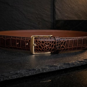 May include: A brown belt with a textured crocodile-embossed pattern and a polished gold-tone metal buckle. The belt features a smooth tan interior lining and a single matching leather keeper loop, showcasing a classic design with visible stitching.