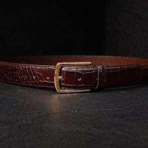 May include: A dark brown belt featuring a textured crocodile-embossed pattern across the strap. The belt is secured with a polished gold-tone metal buckle and a matching leather keeper loop, showcasing a classic design with visible stitching.
