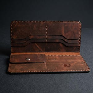 Puede incluir: Una cartera larga de cuero marrón con diseño horizontal que incluye múltiples ranuras para tarjetas, un bolsillo para monedas con cierre de botón metálico y un pequeño logotipo grabado en la parte inferior derecha con costuras.