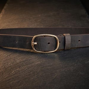 May include: A dark brown leather belt featuring a smooth, polished gold-tone oval buckle. The belt strap has a matte finish with visible adjustment holes and a matching leather keeper loop positioned next to the buckle for a secure fit.