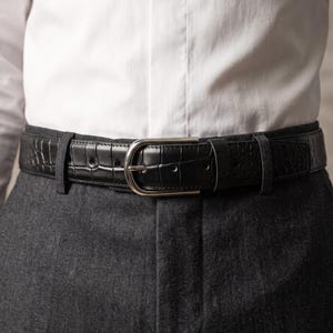 May include: A black leather belt featuring a textured crocodile-embossed pattern and a polished silver-tone metal buckle. The belt is threaded through the belt loops of dark grey trousers, worn below a crisp white button-down shirt with a central placket.