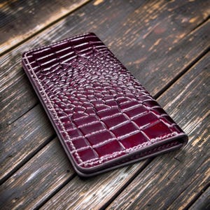 May include: A burgundy leather wallet with a crocodile skin texture. The wallet is rectangular with a stitched border. The surface has a glossy finish, reflecting light. The wallet is placed on a dark wooden surface.