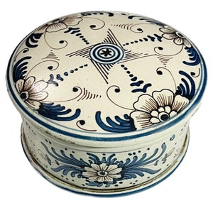 May include: A white ceramic trinket box with a blue and brown floral design. The lid features a central starburst pattern.