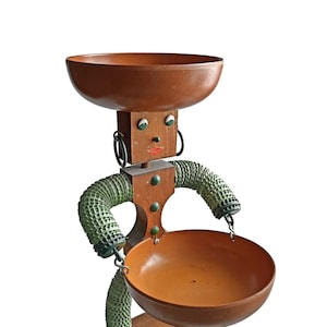 Pode incluir: Uma figura de madeira com forma humana, segurando duas tigelas. A figura tem um rosto castanho com olhos pintados e uma boca vermelha. Os braços da figura são verdes e têm um padrão texturizado. As tigelas são castanhas e de madeira.