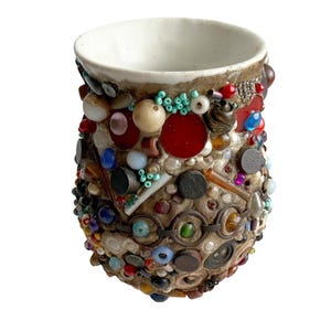 Puede incluir: Una taza artesanal de estilo mosaico con interior blanco y exterior texturizado. El exterior está adornado con una variedad de cuentas coloridas, botones y pequeños elementos decorativos en tonos rojos, azules, verdes y marrones.