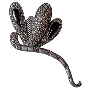 Puede incluir: Un broche con forma de libélula hecho de metal plateado, con incrustaciones de marcasita en las alas y la cola segmentada. El diseño incluye dos piedras negras lisas y pulidas en forma de lágrima engastadas en las alas superiores.