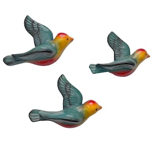 May include: Three ceramic bird figurines in flight. The birds have teal wings, yellow and orange heads, and red bellies. The wings are detailed with etched feather patterns. The birds are set against a white background.