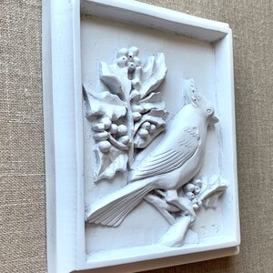 Puede incluir: Placa de pared de cer&aacute;mica blanca con una escultura en relieve de un p&aacute;jaro posado en una rama con bayas de acebo. La placa est&aacute; enmarcada con un borde blanco y firmada "J 79" en la esquina inferior derecha.