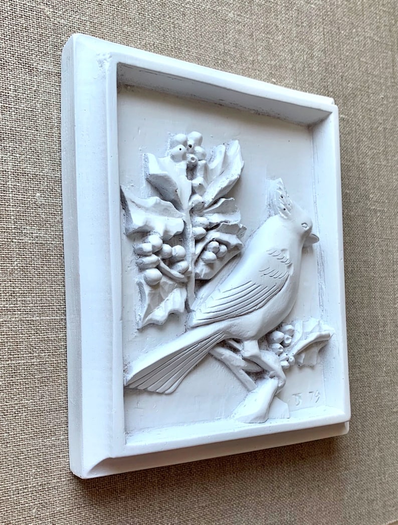 Puede incluir: Placa de pared de cer&aacute;mica blanca con una escultura en relieve de un p&aacute;jaro posado en una rama con bayas de acebo. La placa est&aacute; enmarcada con un borde blanco y firmada "J 79" en la esquina inferior derecha.