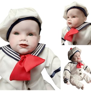 May include: A porcelain doll dressed in a sailor outfit. The doll wears a white sailor suit with navy blue trim, a red neckerchief, and a matching white hat with a navy blue band. The doll has green eyes and rosy cheeks.