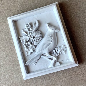 Puede incluir: Decoraci&oacute;n de pared enmarcada en blanco con una escultura en relieve 3D de un cardenal posado en una rama de acebo con bayas. La escultura est&aacute; pintada de blanco y tiene un aspecto vintage. La esquina inferior derecha del marco est&aacute; firmada con "D 79".