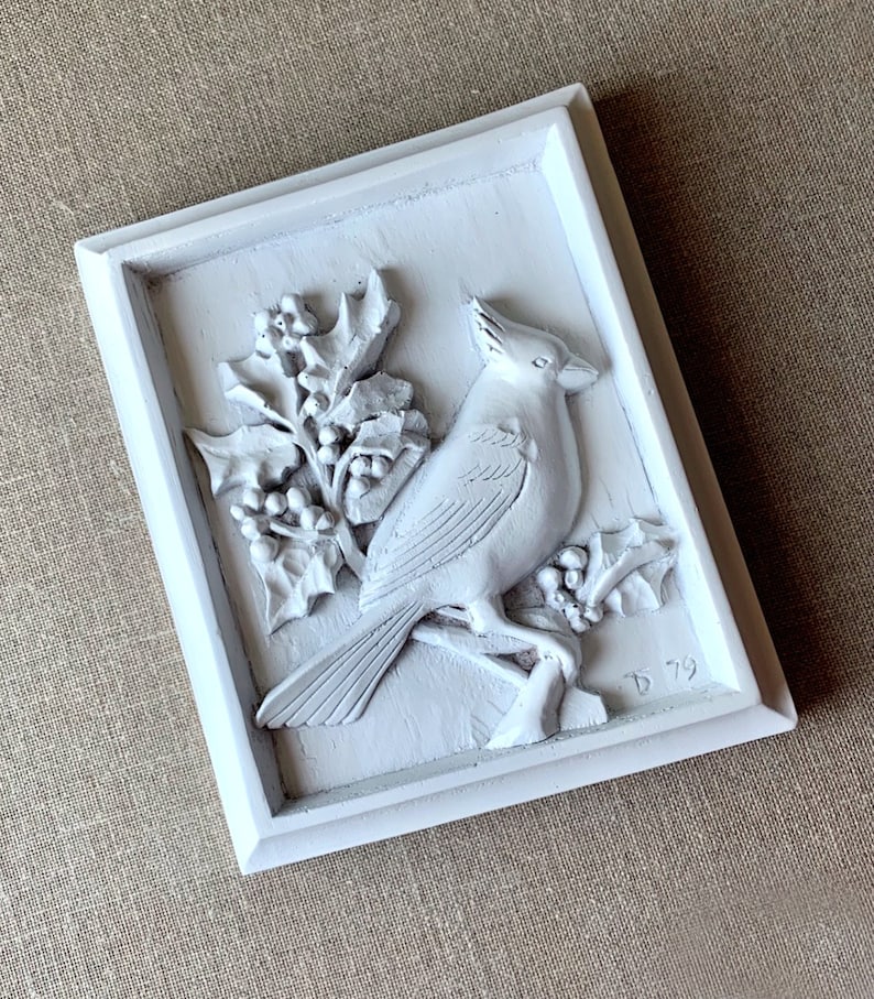 Puede incluir: Decoraci&oacute;n de pared enmarcada en blanco con una escultura en relieve 3D de un cardenal posado en una rama de acebo con bayas. La escultura est&aacute; pintada de blanco y tiene un aspecto vintage. La esquina inferior derecha del marco est&aacute; firmada con "D 79".
