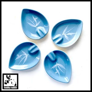 May include: Four light blue ceramic ashtrays with a white floral design. Each ashtray has a rounded, teardrop shape with a small indentation for cigarettes.