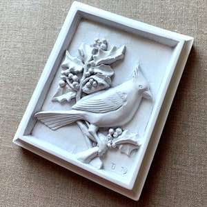 Puede incluir: Placa de pared de cer&aacute;mica blanca con una escultura en relieve 3D de un cardenal posado en una rama de acebo con bayas rojas. La placa est&aacute; enmarcada con un borde blanco y tiene la inscripci&oacute;n "B 79" en la esquina inferior derecha.