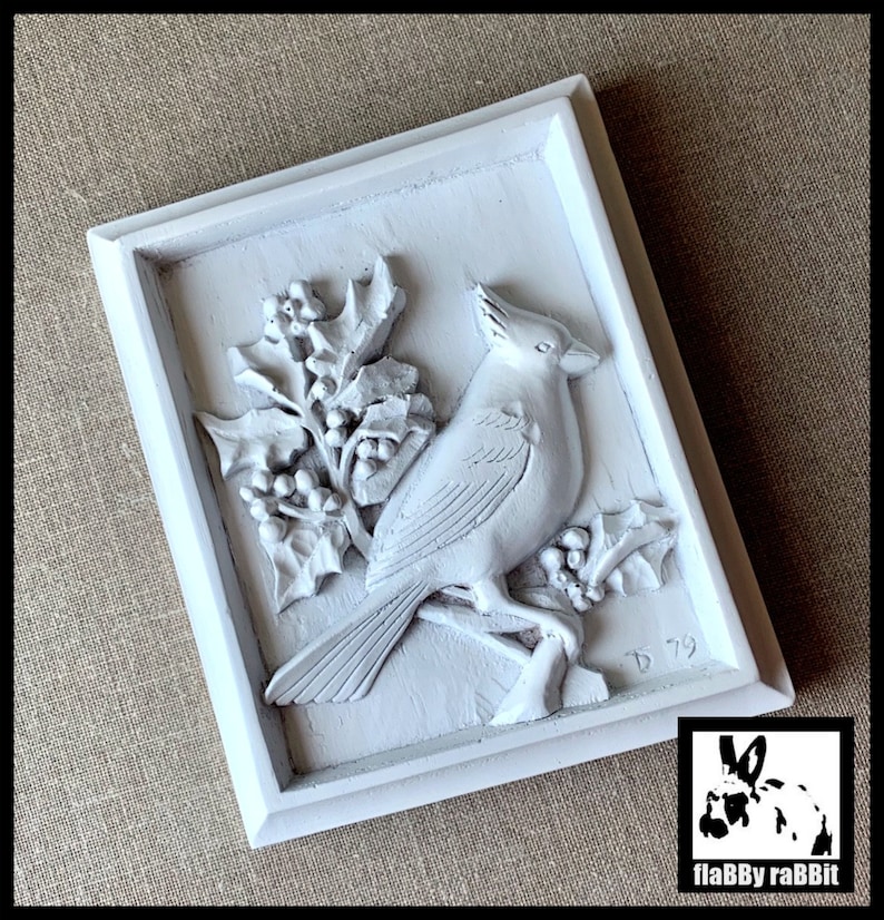 Puede incluir: Decoraci&oacute;n de pared enmarcada en blanco con una escultura en relieve 3D de un cardenal posado en una rama de acebo con bayas rojas. La escultura est&aacute; firmada "D 79".
