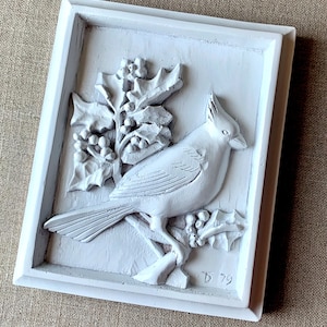 Puede incluir: Placa de pared de cer&aacute;mica blanca con un dise&ntilde;o en relieve de un cardenal posado en una rama de acebo con bayas rojas. La placa est&aacute; enmarcada en un marco blanco y tiene el n&uacute;mero "D 79" inscrito en la esquina inferior derecha.