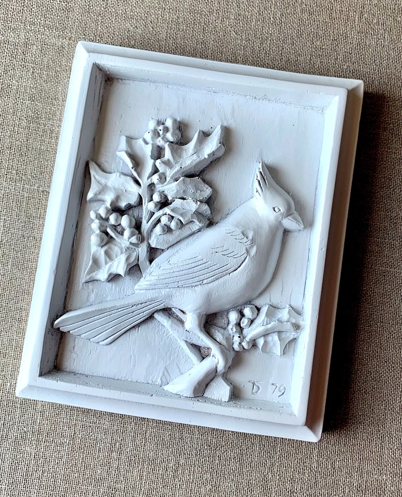 Puede incluir: Placa de pared de cer&aacute;mica blanca con un dise&ntilde;o en relieve de un cardenal posado en una rama de acebo con bayas rojas. La placa est&aacute; enmarcada en un marco blanco y tiene el n&uacute;mero "D 79" inscrito en la esquina inferior derecha.