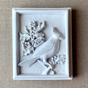 Puede incluir: Placa de madera pintada de blanco con una escultura en relieve de un cardenal posado en una rama de acebo con bayas. La placa est&aacute; firmada "D 79" en la esquina inferior derecha.