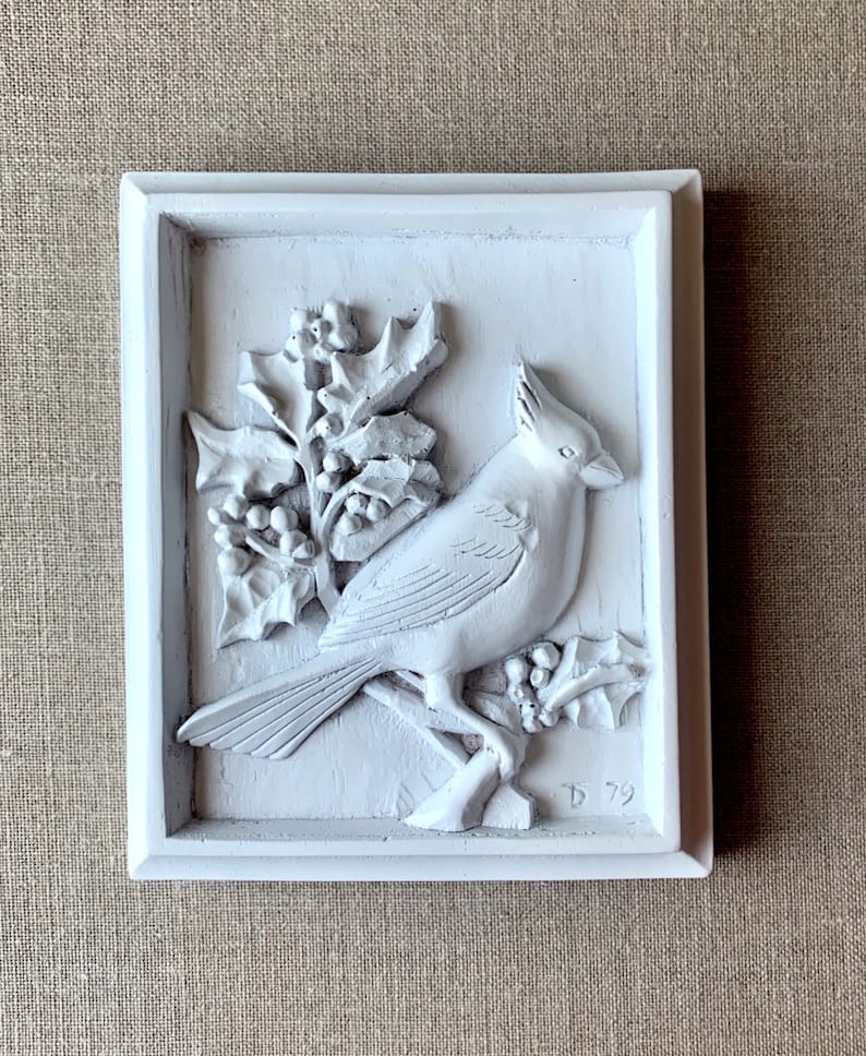Puede incluir: Placa de madera pintada de blanco con una escultura en relieve de un cardenal posado en una rama de acebo con bayas. La placa est&aacute; firmada "D 79" en la esquina inferior derecha.
