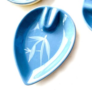 May include: A light blue ceramic ashtray with a white stylised bamboo design. The ashtray is shaped like a leaf and has a small indentation for cigarettes.