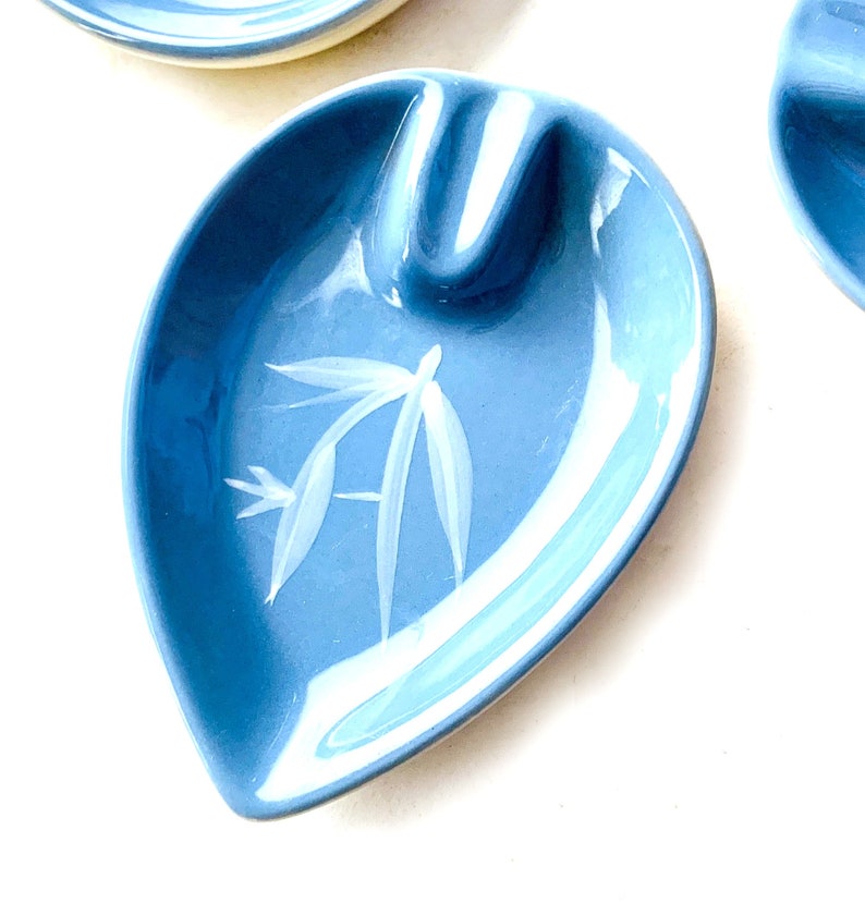 May include: A light blue ceramic ashtray with a white stylised bamboo design. The ashtray is shaped like a leaf and has a small indentation for cigarettes.
