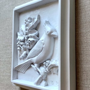 Puede incluir: Placa de pared de cer&aacute;mica blanca con un dise&ntilde;o en relieve de un cardenal posado en una rama de acebo. La placa est&aacute; enmarcada con un borde blanco y tiene la inscripci&oacute;n "D 74" en la esquina inferior derecha.
