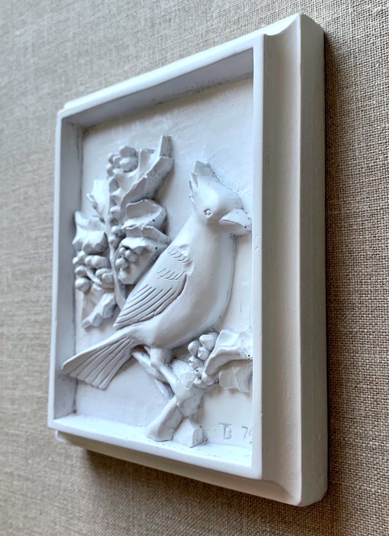 Puede incluir: Placa de pared de cer&aacute;mica blanca con un dise&ntilde;o en relieve de un cardenal posado en una rama de acebo. La placa est&aacute; enmarcada con un borde blanco y tiene la inscripci&oacute;n "D 74" en la esquina inferior derecha.