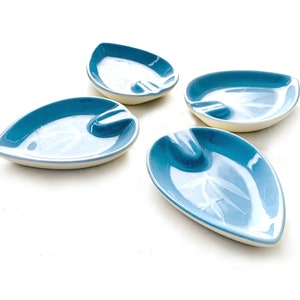 May include: Four blue ceramic leaf-shaped ashtrays with a white rim and a white design on the surface.