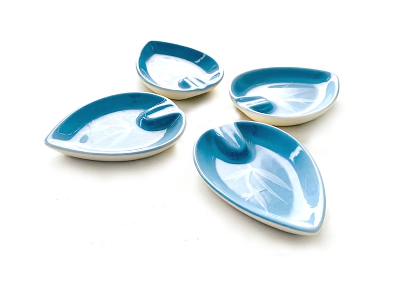 May include: Four blue ceramic leaf-shaped ashtrays with a white rim and a white design on the surface.