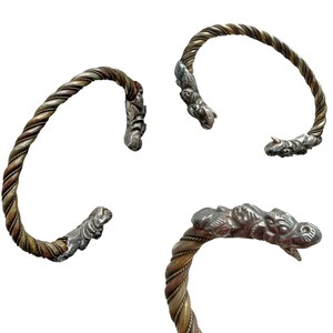 May include: Three silver and bronze-toned twisted metal bracelets. Each bracelet features a detailed animal head design at the ends. The bracelets are displayed against a white background.