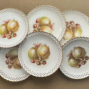 Könnte beinhalten: Sechs weiße Porzellan-Dessertteller mit goldfarbenem Rand und einem filigranen durchbrochenen Design. Jeder Teller zeigt ein handgemaltes Design aus zwei Pfirsichen und Haselnüssen.