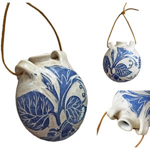 Op de afbeelding: Handgemaakte keramische hanger met een blauw en wit bloemmotief. De hanger is rond met een kleine tuit en twee handvatten. Hij hangt aan een bruin leren koord.
