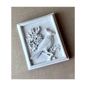 Puede incluir: Placa de madera pintada de blanco con una escultura en relieve de un cardenal posado en una rama de acebo. La placa est&aacute; enmarcada con un borde blanco y tiene la fecha "D 79" inscrita en la esquina inferior derecha.