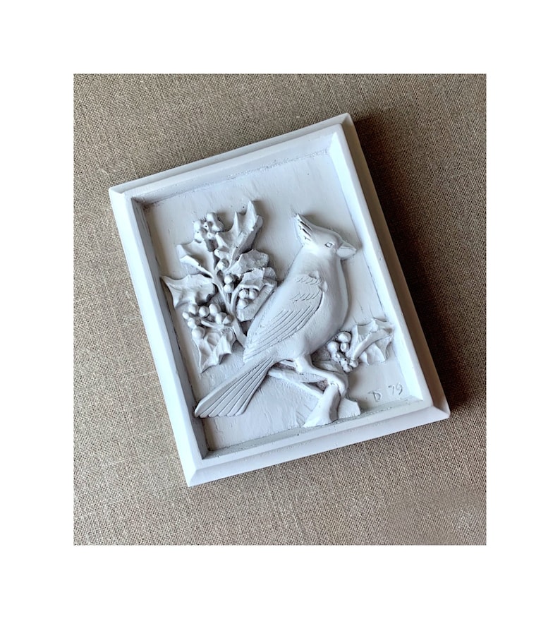 Puede incluir: Placa de madera pintada de blanco con una escultura en relieve de un cardenal posado en una rama de acebo. La placa est&aacute; enmarcada con un borde blanco y tiene la fecha "D 79" inscrita en la esquina inferior derecha.