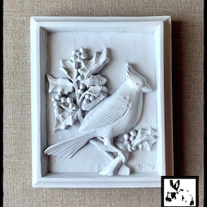 Puede incluir: Placa de pared pintada de blanco con una escultura en relieve 3D de un cardenal posado en una rama de acebo con bayas. La placa tiene un marco blanco y la fecha '79' es visible en la esquina inferior derecha.