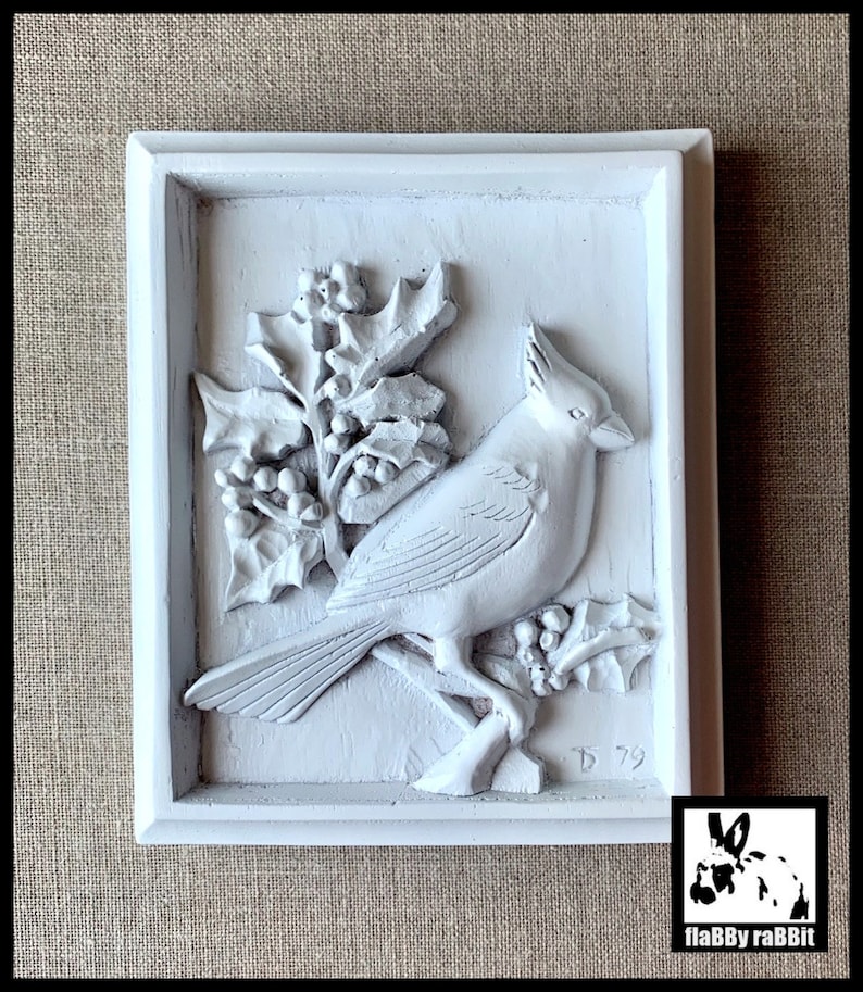 Puede incluir: Placa de pared pintada de blanco con una escultura en relieve 3D de un cardenal posado en una rama de acebo con bayas. La placa tiene un marco blanco y la fecha '79' es visible en la esquina inferior derecha.