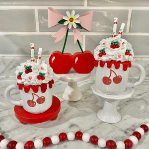 May include: A decorative arrangement featuring cherry-themed items. Includes two mugs with cherry designs and whipped cream toppings, a cherry ornament, and a beaded garland. The mugs sit on red and white pedestals, with a pink bow and daisy accent.
