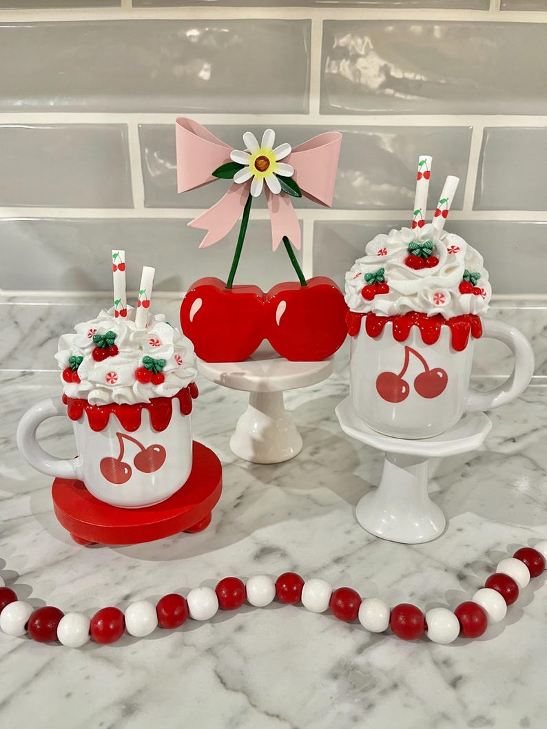 May include: A decorative arrangement featuring cherry-themed items. Includes two mugs with cherry designs and whipped cream toppings, a cherry ornament, and a beaded garland. The mugs sit on red and white pedestals, with a pink bow and daisy accent.