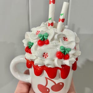 May include: A white ceramic mug with a cherry design, topped with whipped cream, red cherry accents, and two straws. The mug has a red cherry design on the front and red drips around the rim. The straws are white with red cherry illustrations.