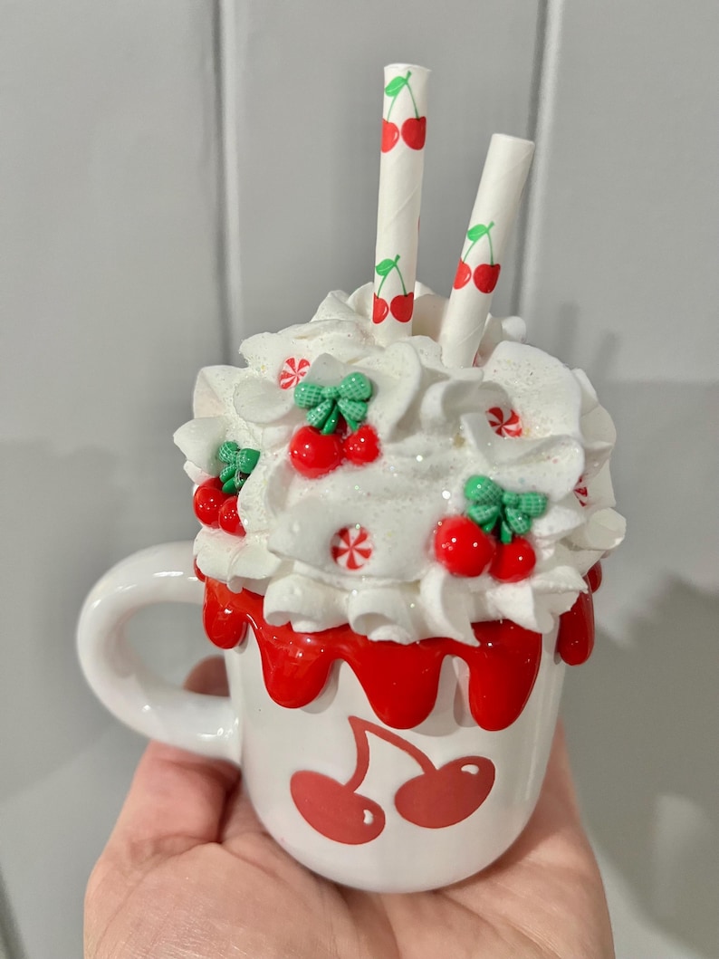 May include: A white ceramic mug with a cherry design, topped with whipped cream, red cherry accents, and two straws. The mug has a red cherry design on the front and red drips around the rim. The straws are white with red cherry illustrations.